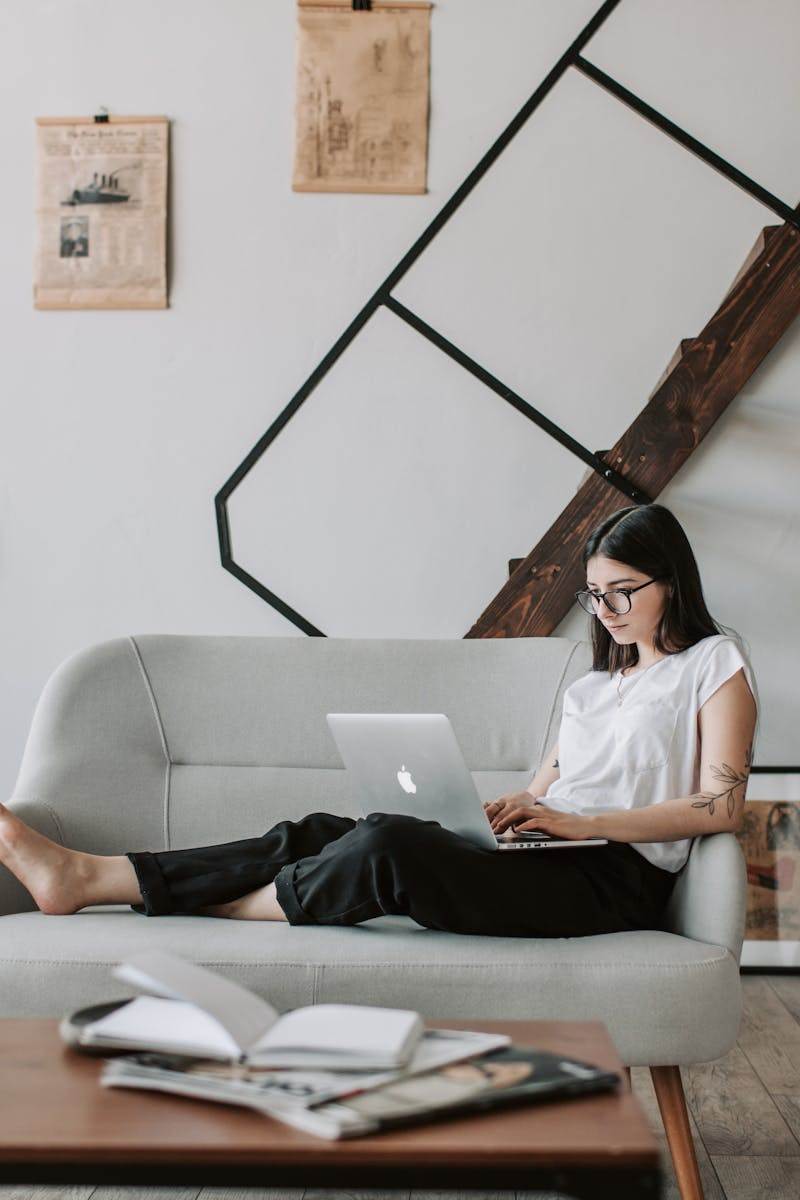 Positive female with tattooed arms wearing casual clothes and eyeglasses reading a humbee newsletter while sitting barefoot on comfortable sofa behind table with books and magazines in modern apartment with minimalist interior in eco style during free time